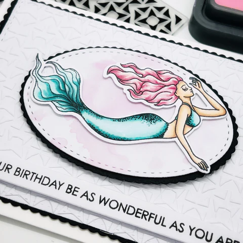 close up of the mermaid birthday card, embossed with dry embossing in triangular style,  with black scallop border. a oval black scallop to border the stitched oval die cut that showcases the mermaid image, with coral pink hair and aqua green on body and bikini top. with stamped sentiment in black 'may your birthday be as wonderful as you are'.