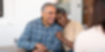 Smiling couple sitting together on a couch, woman resting her head on man's shoulder. He wears a blue plaid shirt, she wears a beige sweater. Cozy room.