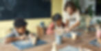 Children sculpt clay at a wooden table, guided by a standing woman in an apron. Background: a blackboard and yellow wall. Creative mood.