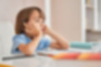Child in blue shirt, holding a pencil, daydreams at a desk with open notebook. Books and soft focus background suggest a study environment.