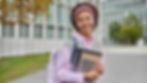 Young woman with curly hair and pink headband smiles, holding books and tablet. She's outside a modern building, wearing a pink shirt and backpack.