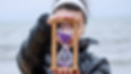 Person in a black jacket holds a wooden hourglass with purple sand against a blurry ocean background. The mood is contemplative.