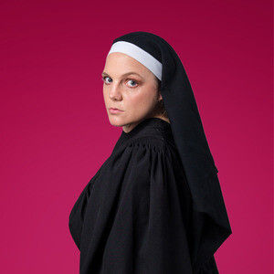 Woman in a black nun’s habit and white coif looks at the camera against a bright pink background.
