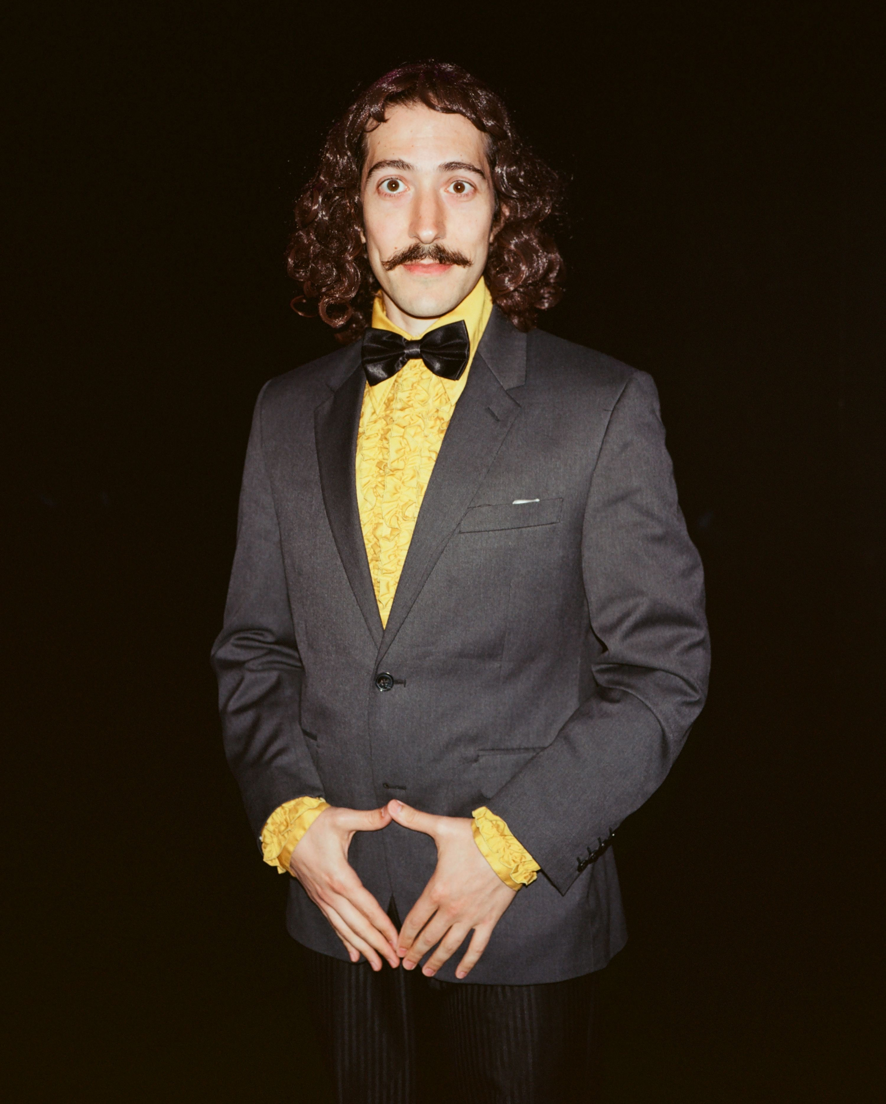 Man in a gray suit with a yellow ruffled shirt and bow tie, posing against a black background with hands forming a heart.
