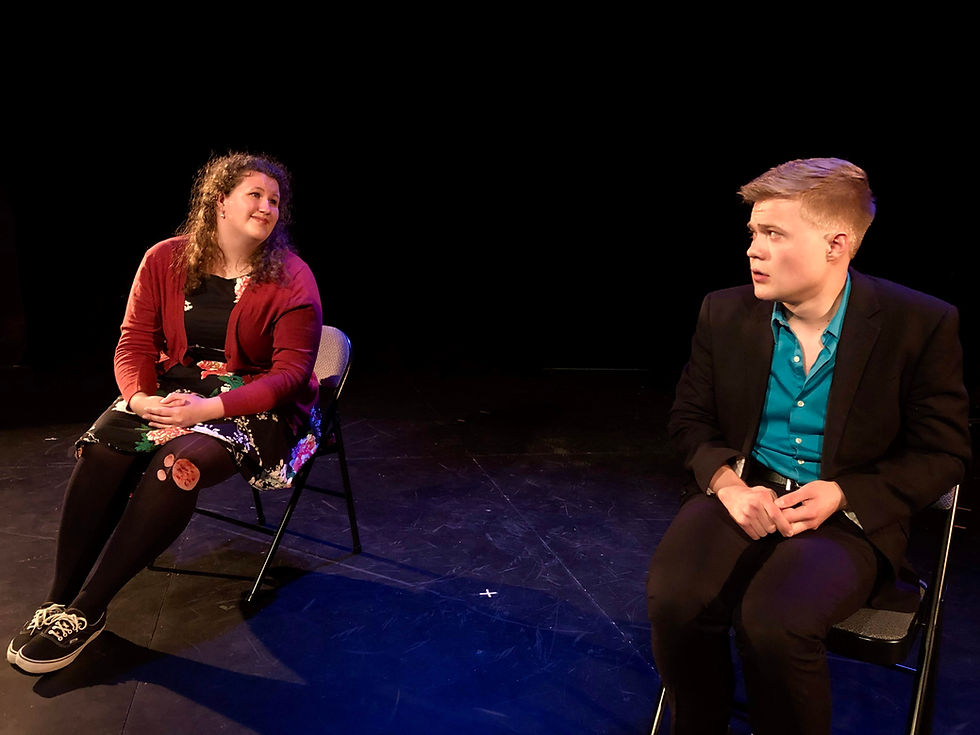 Two seated performers on a dark stage, a woman in red smiling toward a man in a teal shirt and black blazer, both thoughtful. Minnesota Fringe Artists Rachel Ropella and Timothy Kelly.