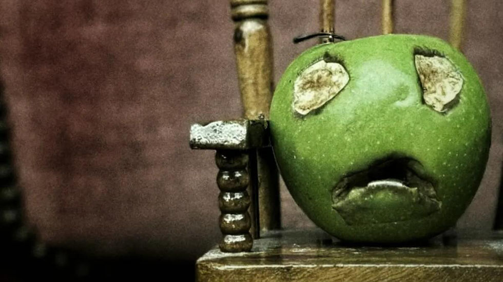 Green apple carved with a sad face sits on a rustic wooden chair against a dim brown background.