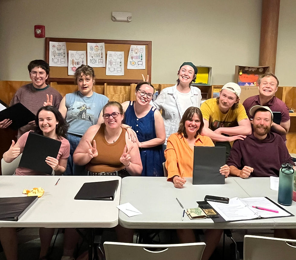 Smiling group of nine in a classroom, posing with black folders and peace signs at a table with papers and supplies. Minnesota Fringe Artists Melancholics Anonymous.