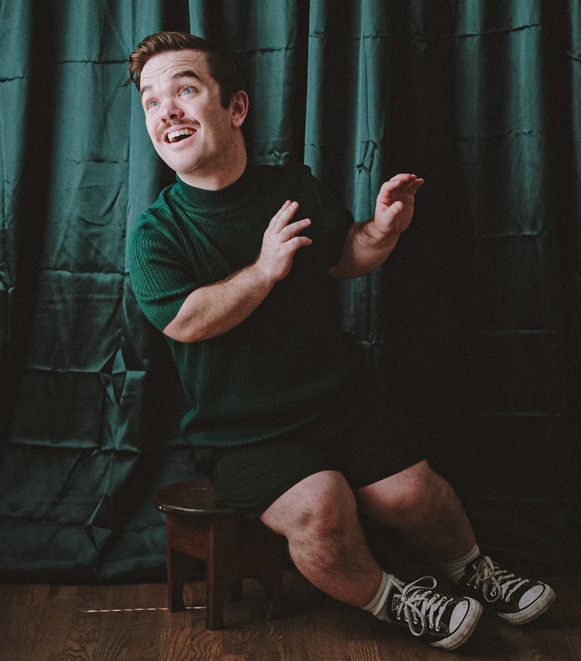 Man in a green sweater and shorts sits on a stool before a teal curtain, smiling with a surprised, playful pose.