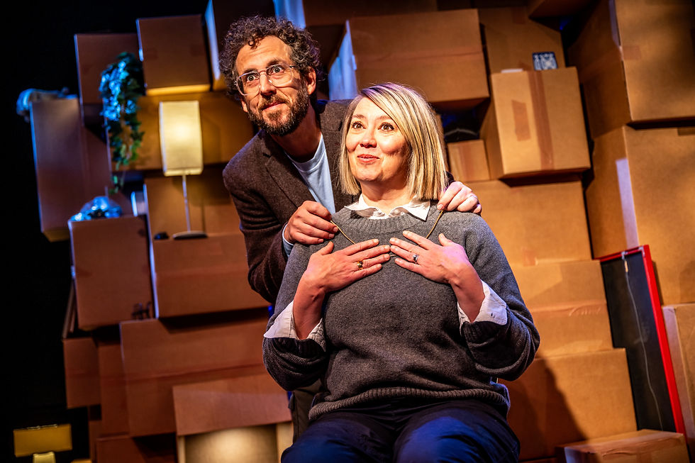 Two people pose on a warm-lit stage of stacked cardboard boxes, the man standing behind the seated woman with hands on her shoulders.