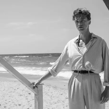 Un homme en chemise d'été se tient à une rambarde d'un cabanon de plage. Il est sur le sable, dos à la mer, le regard vide.