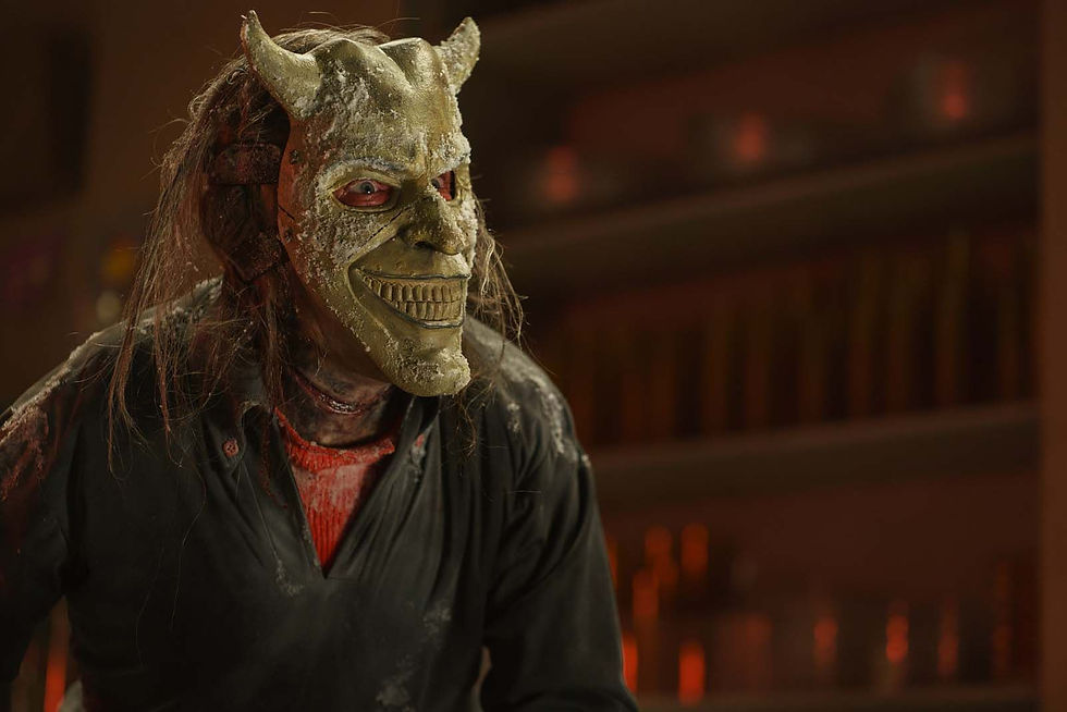 Un homme avec des vêtements et des cheveux sales. Il porte un masque gris en forme de démon et avec un sourire menaçant.