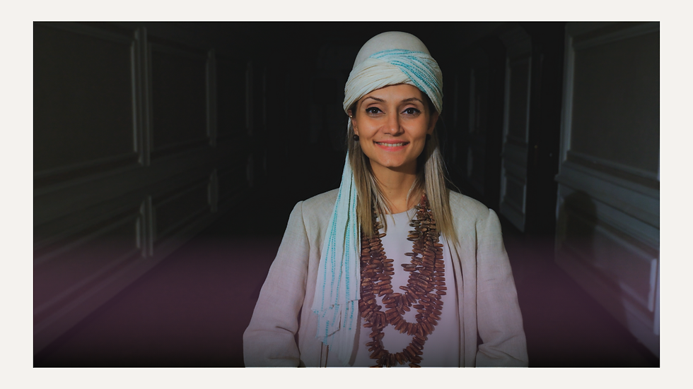 Geleneksel görünümlü beyaz bir başörtü ve uzun boncuklu kolyeler takan, beyaz ceket giymiş genç bir kadın, loş bir koridorda gülümseyerek poz veriyor.