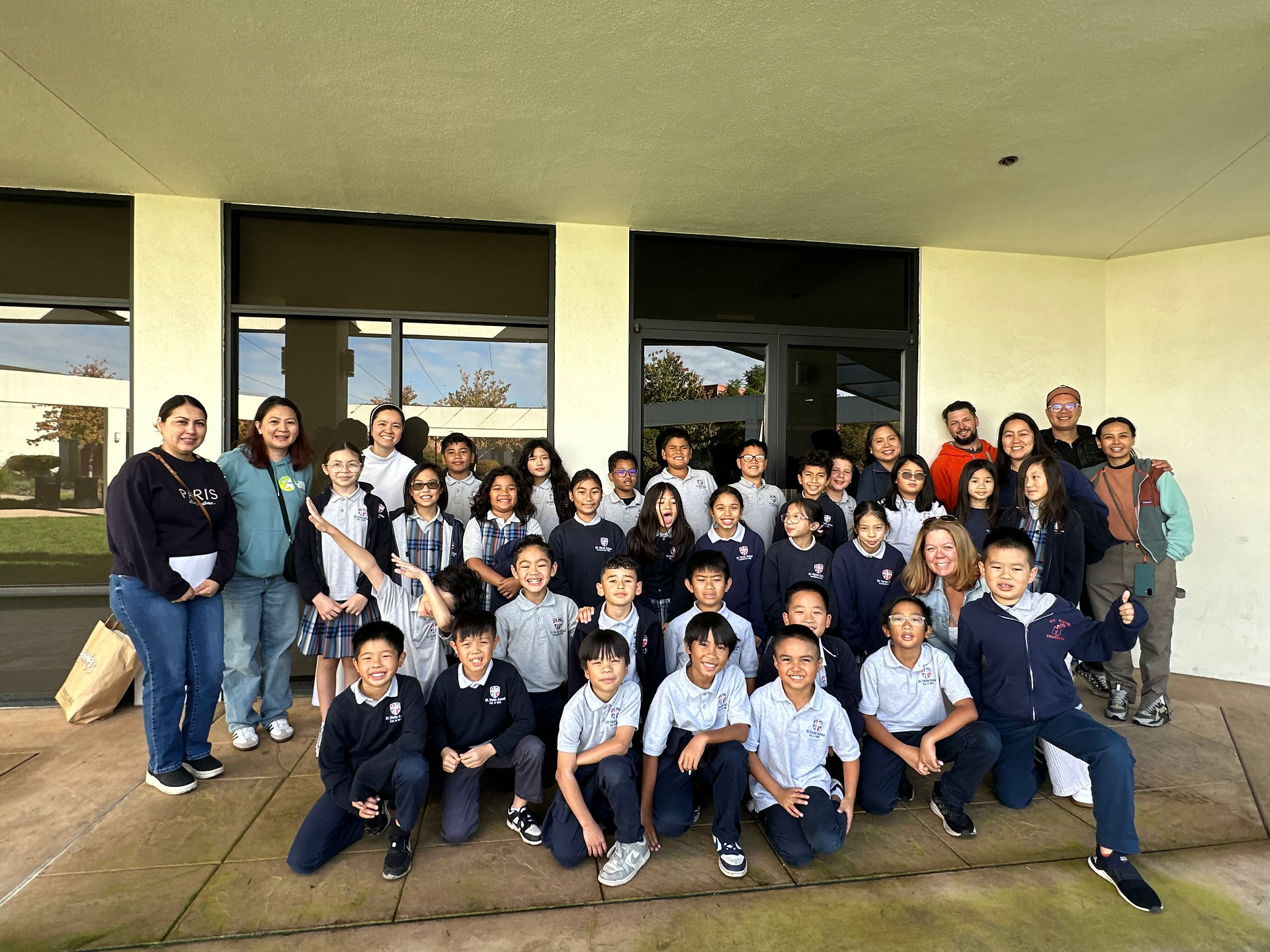 Students, teachers, and several parent chaperones pose together for a cheerful group photo outside the school building.