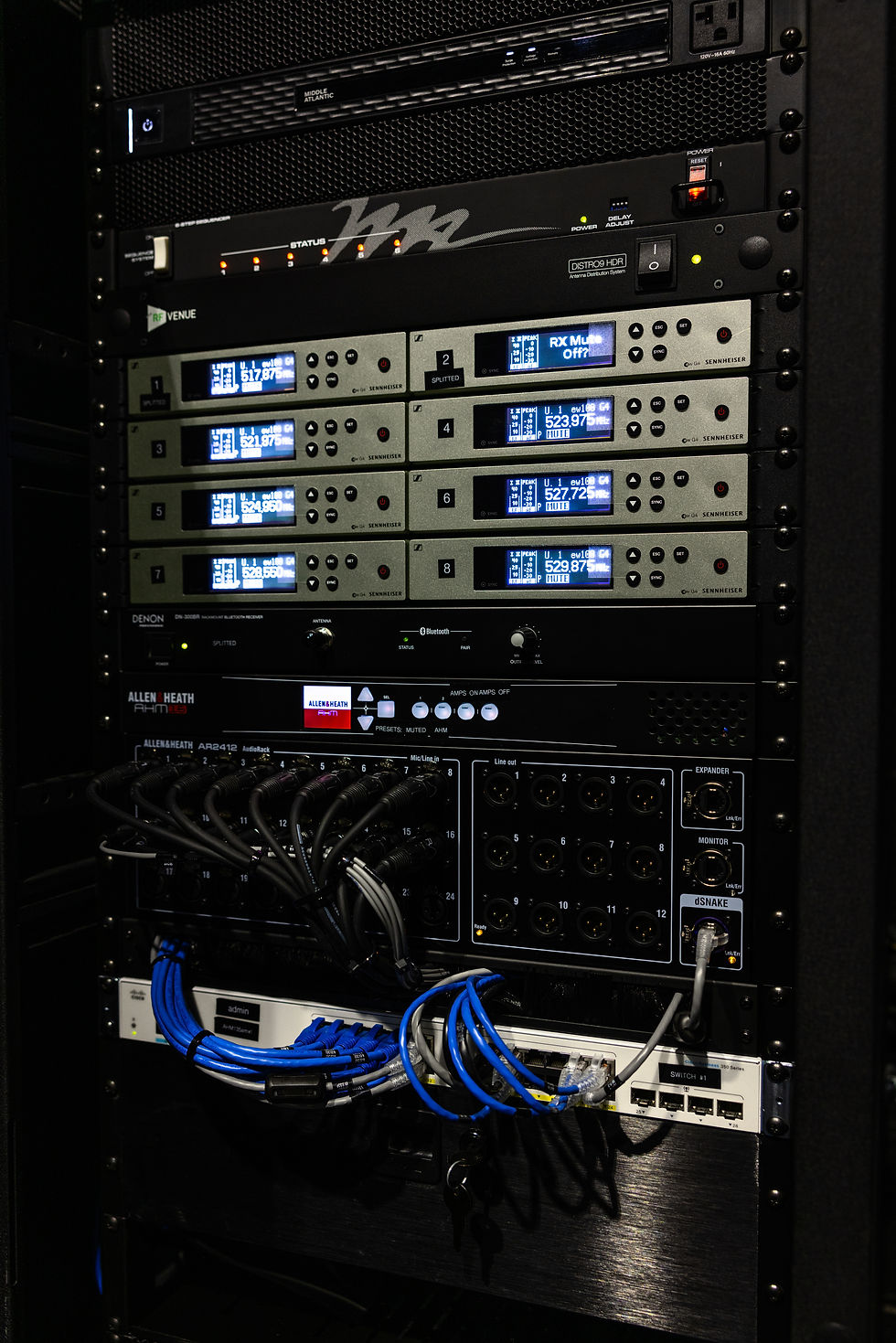 Professional AV equipment rack featuring Sennheiser wireless microphone receivers, an Allen & Heath audio processor, and organized network cabling in a commercial audiovisual installation.