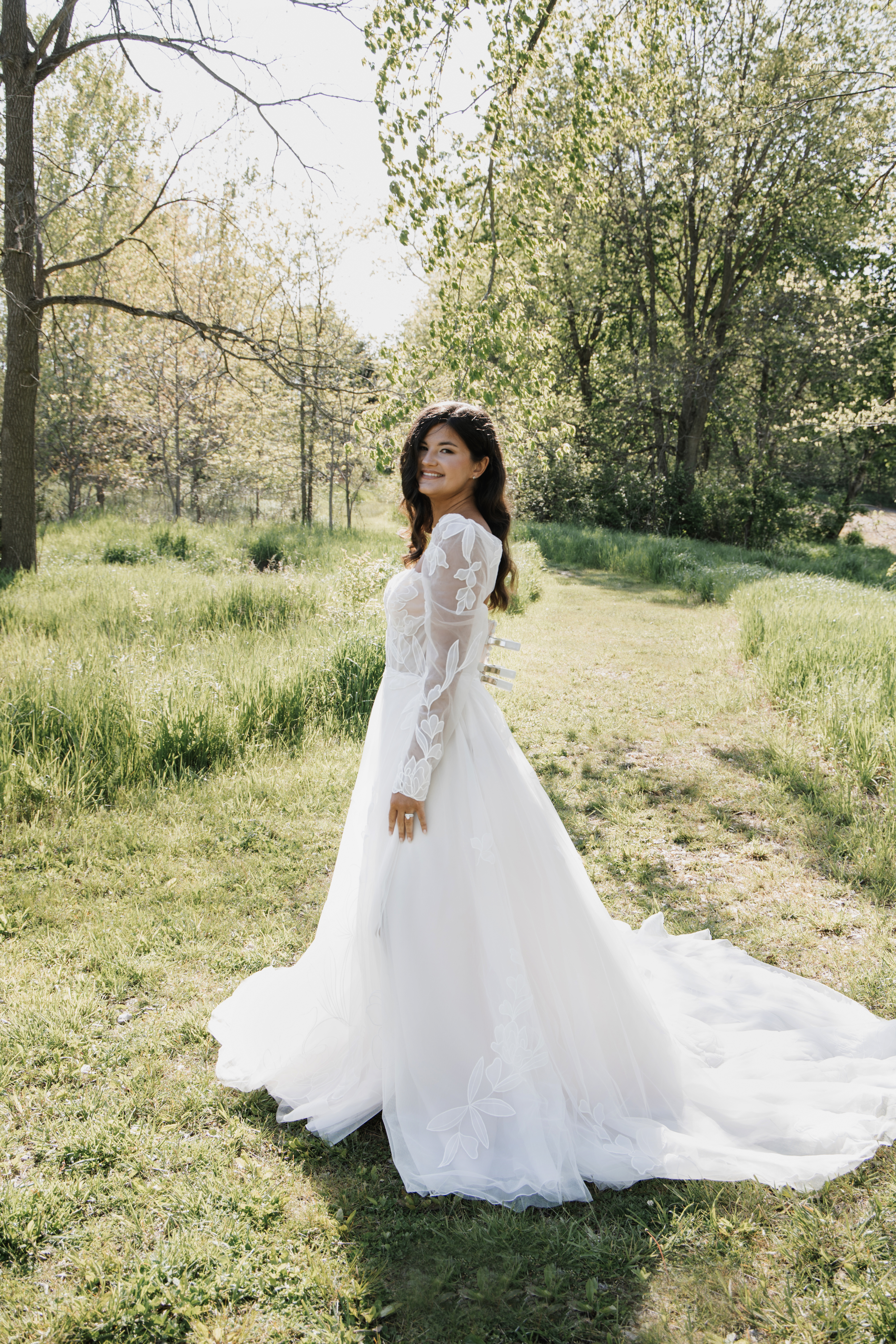 Smiling bride in a long white lace wedding gown stands on a grassy path in a sunlit wooded meadow.