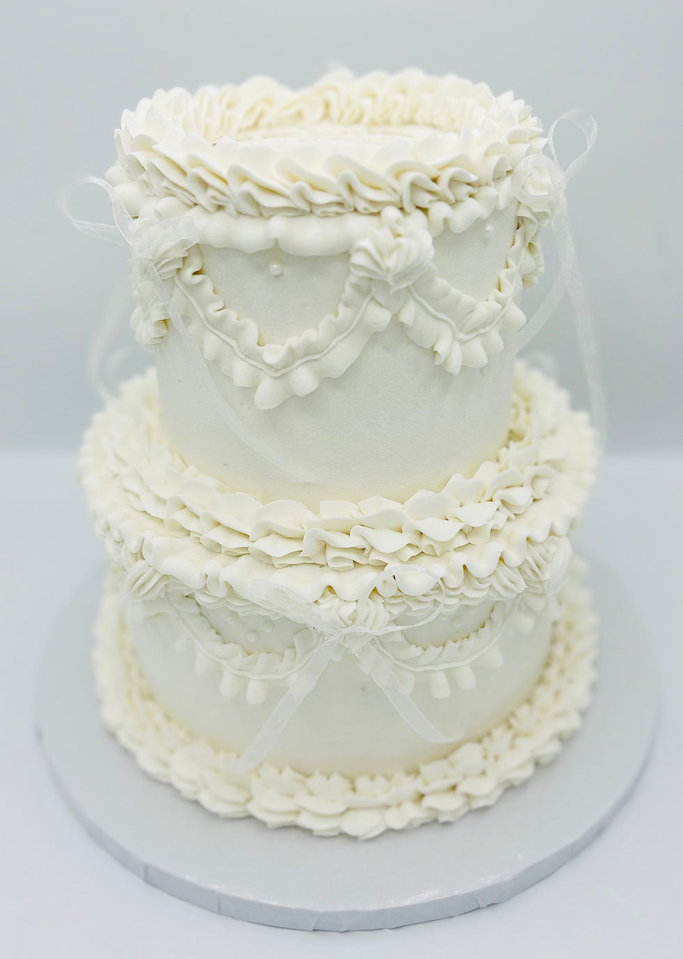 Ornate white tiered cake with ruffled icing and sheer ribbon details on a white stand against a pale background