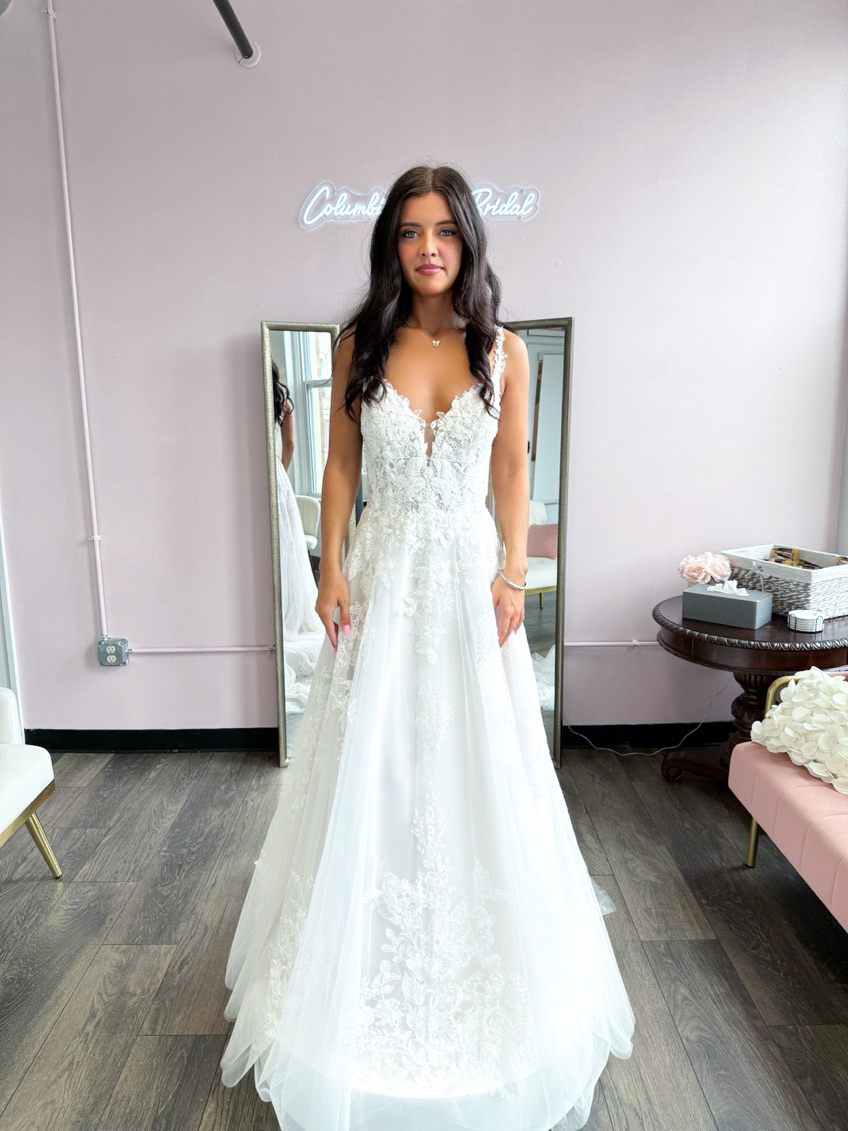 Woman in a white lace wedding gown stands in a bridal shop, with a neon Columbia Bridal sign behind her and mirrors nearby.
