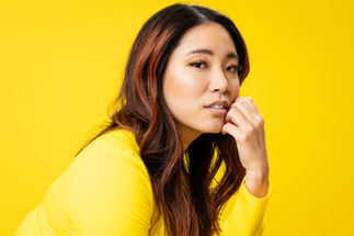 Close up photo portrait of a beautiful young asian woman wearing a yellow sweater in front of a yellow backdrop. She has reddish-orange streaks in her black hair. Her left hand rests gently on her cheek.