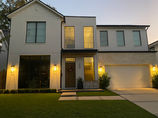 A modern two-story house at dusk with large, sleek windows that reflect soft evening light. The windows, which add to the home’s contemporary design, are spotless and gleaming, suggesting recent professional window cleaning. Warm exterior lights on both levels illuminate the white facade, stone accents, and a well-manicured lawn, creating a welcoming atmosphere.