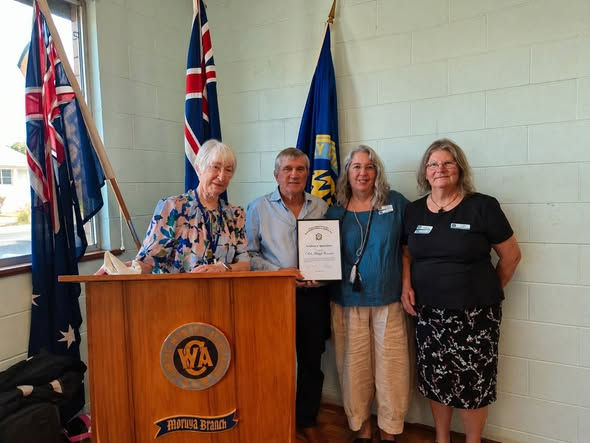 Four people stand smiling beside a wooden podium in a room with flags. One holds a certificate. The mood is formal and celebratory.