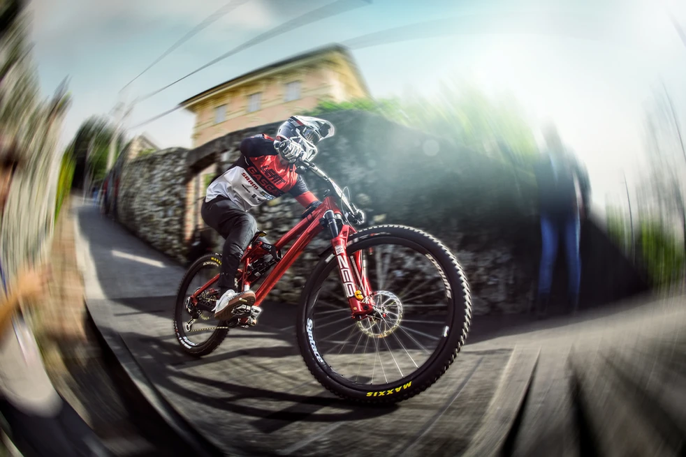 A cyclist speeds down the narrow wooden path of the Cerro Abajo course in Genoa, captured in a high-energy moment with motion blur. The image highlights the thrilling nature of Red Bull Downhill, blending lifestyle and sport photography to capture the essence of competitive cycling.