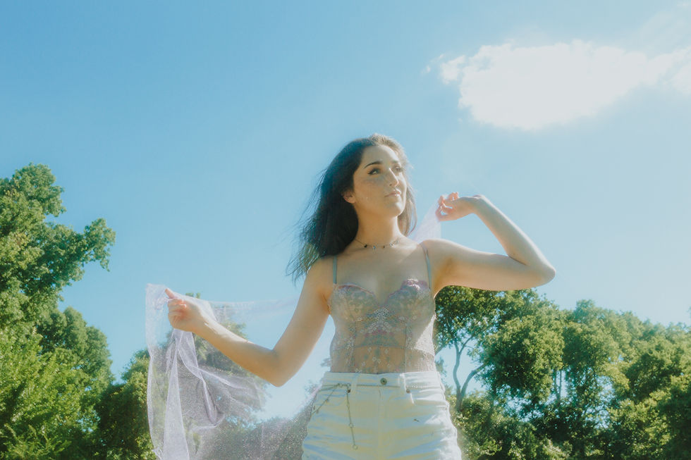 Recording artist Emma Rowley poses under a blue sky in front of green summer trees. The sun shines down as she poses with a piece of shimmering lavender fabric, her hair blowing in the breeze as she looks off into the distance.