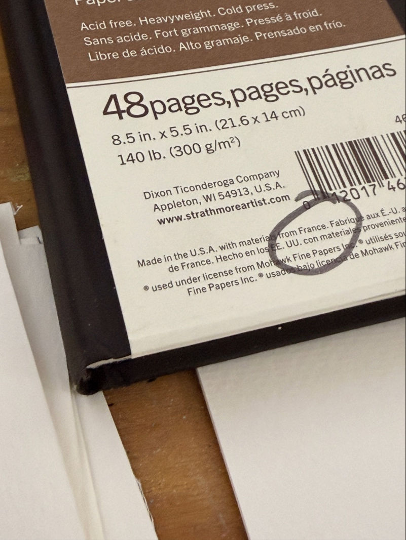Close-up of a sketchbook pad label showing 48 pages, 8.5 x 5.5 in, 140 lb cold press paper on a table.