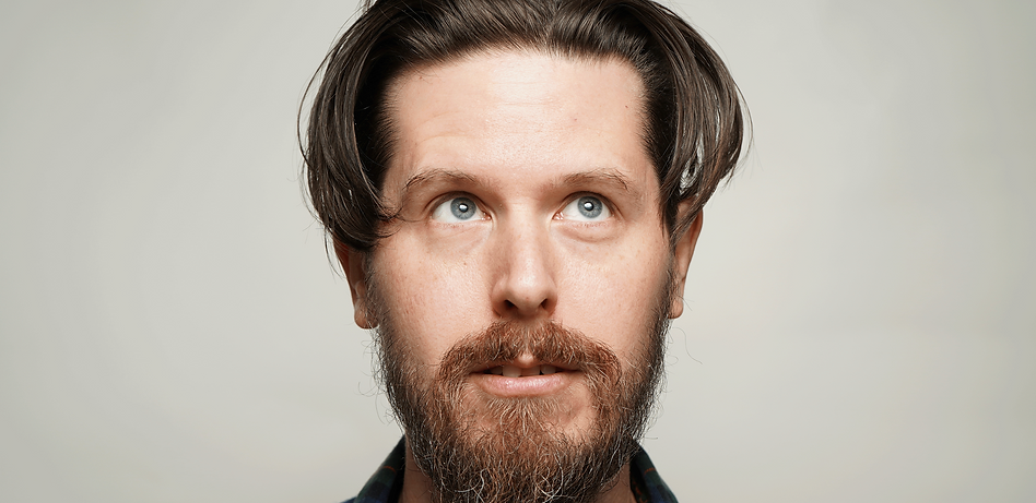 Greyscale headshot of a young white man with medium length dakr hair and short beard looking up.
