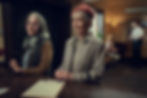 Helen Mirren as Elizabeth in a disguise with Celia Imrie as Joyce standing at a desk with smirks on their face