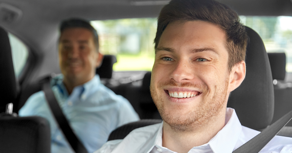 Homem sorridente no banco dianteiro de um carro, com outro passageiro desfocado atrás, clima alegre.