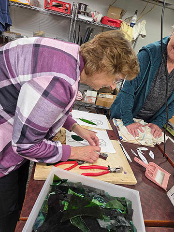 Two people work on a stained glass project in a workshop. One cuts glass, while colorful pieces and tools are spread on a table.