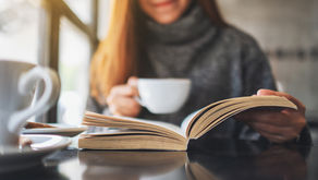 woman in a gray sweater holding a white cup reading a book next to a large window