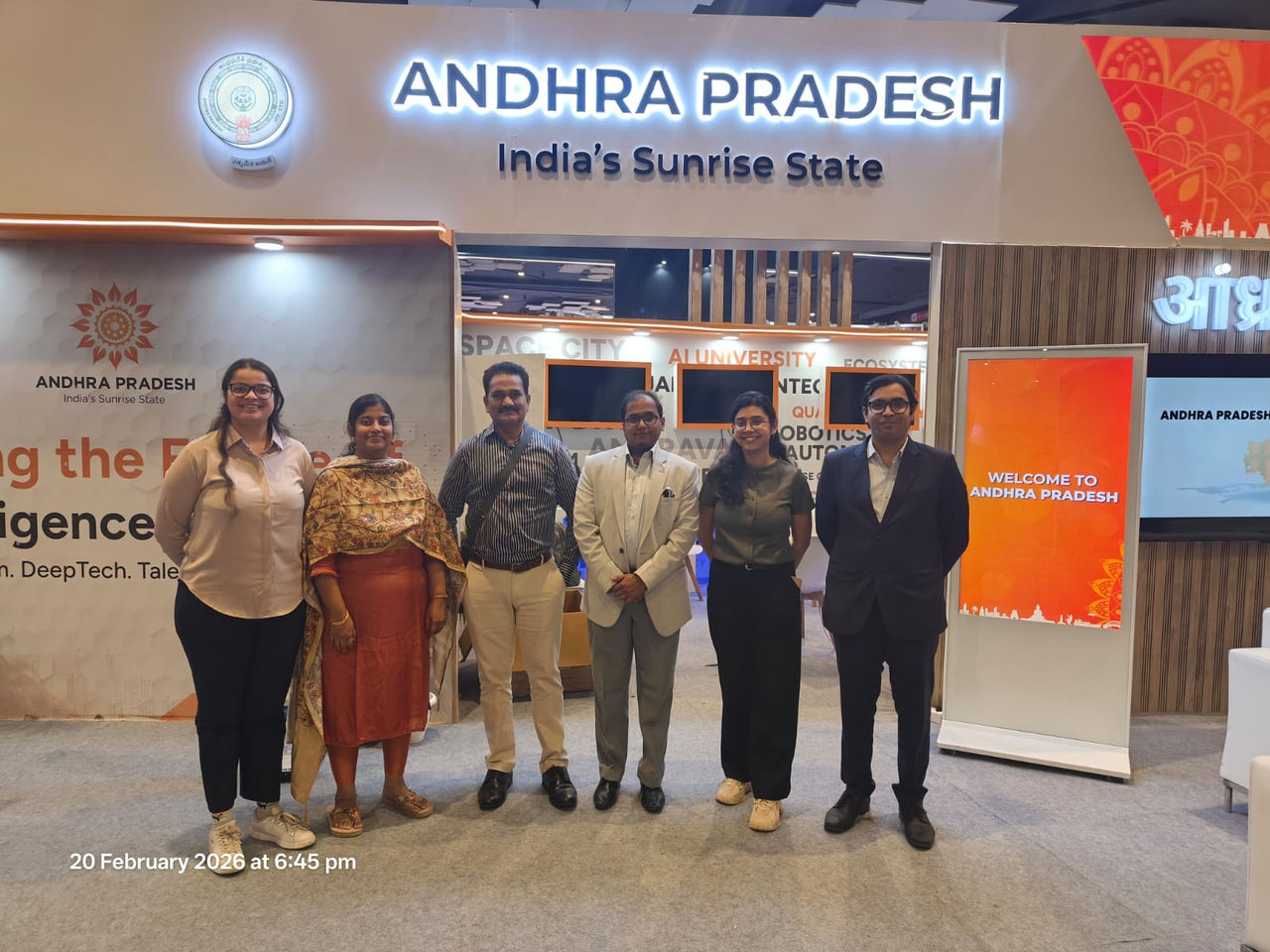 Six Mandelbulb team members standing shoulder to shoulder at an exhibition stand, the lit backdrop of a state government technology pavilion and its state tagline filling the wall behind them.