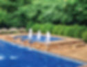 Water fountains in a swimming pool, surrounded by stone and greenery.