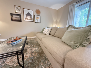 Cushioned sofa with throw pillows and a glass coffee table beside a curtained window, furnished 1 bedroom rental