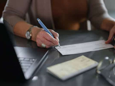 Person writing a bank check with a pen on a desk next to a laptop and checkbook, demonstrating how to write a check in the USA.