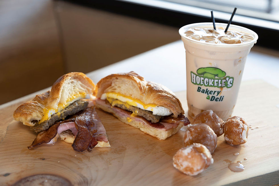 Croissant sandwich with bacon, sausage, and egg on a wooden board, iced coffee with Hoeckele’s Bakery & Deli logo, and powdered donuts.