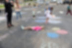 Kids and Misty Brown play on a chalk-decorated parking lot at the 573 Chalk Art Festival. A girl lies on the ground among colorful balloon drawings, creating a fun, lively scene.