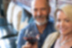 Rob Ray with a beard holds a glass of red wine, smiling beside a Laurie Everett-Ray. They're seated on a bench with cushions and a brick wall backdrop.