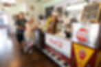 Katey and David John owner of Num Nums stand in a vintage soda shop with Coca-Cola decor, leaning against a counter. Menu board and snacks visible; casual, nostalgic mood.