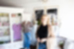 Kim Smith owner of Miss Kim & Co smiling in chic room with wardrobe displaying a purple dress and outfit. Black-and-white photos decorate the wall, creating a cozy vibe.