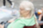 Elderly woman with braided hair sits outdoors in a green shirt, facing right. Blurred background shows pavement and colorful items. A 573 Chalk Art Festival.