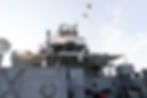 Battleship bridge with flags against a clear sky. Gray metal structures and a lion emblem visible. Tents shade parts of the deck.