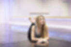Skyler Gollaher of Slay Dance Centre in Bonne Terre, Missouri with long hair, wearing a tank top and leggings, lies on a glossy dance studio floor with mirrored walls, appearing relaxed.