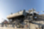 Grey naval ship with a small boat labeled "St 325-1" on deck. Clear blue sky, colorful signal flags. Calm and sunny.