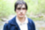Wet-haired Jack Buerck in a navy coat stands in a lush, green forest by a river. Their expression is serious, with a soft background focus. Perryville, Missouri
