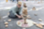 A child named Garett wearing a cap kneels on asphalt, stacking rocks in a tower at the 573 Chalk Art Festival. 573 Magazine circles are around. Others are blurred in the background.