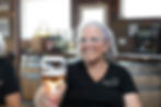 Steffie Littlefield, owner of Edg Clif Winery holding glass of wine in a rustic setting with wine bottles in background. She's wearing glasses and a black shirt.