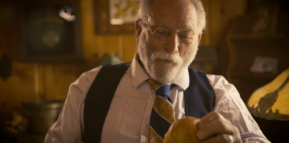 Mickey Hertwick of Marquand Missouri, Elderly man with glasses inspects a tennis ball in a cozy, warmly-lit room. He's wearing a striped shirt, patterned tie, and suspenders.