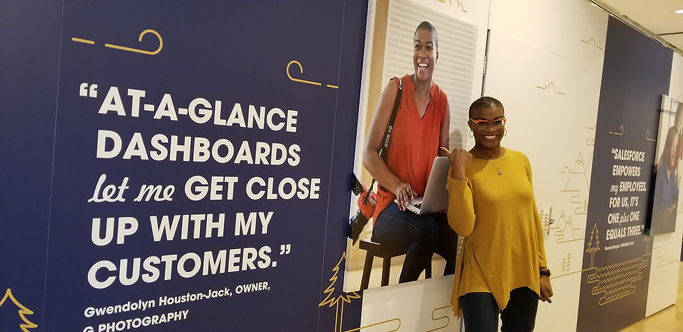 Gwendolyn Houston-Jack a Black woman small business owner poses in front of her Salesforce billboard at Salesforce Dreamforce 2019. She's wearing a marigold yellow top and dark blue jeans with yellow and orange glasses.